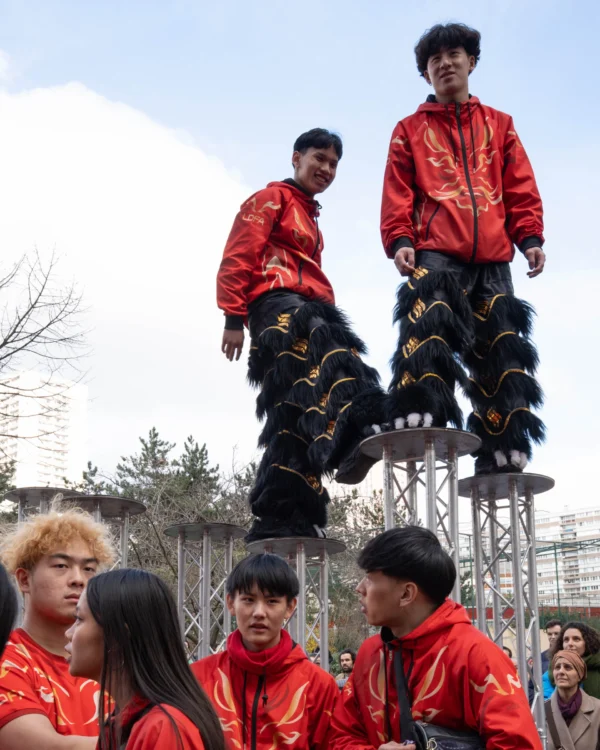Alice · 01/03/2026 · 4017 x 5021 Cette photographie capture un groupe de jeunes en tenues rouges participant à une activité acrobatique ou sportive. Le sujet principal est les trois individus en haut d’une structure pyramidale, portant des chaussures à crampons.La composition montre un cadrage dynamique, avec un premier plan occupé par des spectateurs attentifs. La lumière est naturelle, légèrement voilée par un ciel nuageux. Les couleurs dominantes sont le rouge vif des vêtements et le gris des bâtiments en arrière-plan.L’ambiance est énergique et festive, évoquant une démonstration de force et de coordination dans un contexte probablement traditionnel ou culturel.