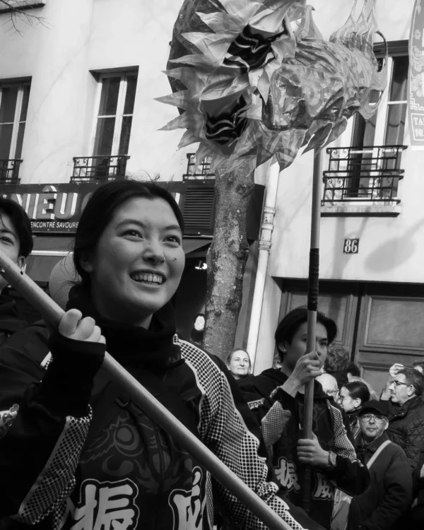 Alice · 1276 x 1595 Cette photographie en noir et blanc met en scène une femme souriante, sujet principal, tenant une perche avec un dragon décoratif. La composition montre une foule en arrière-plan, suggérant une fête ou une procession. Le cadrage est serré sur la femme, avec une légère inclinaison vers le haut pour accentuer le dragon.La lumière semble naturelle, avec des contrastes marqués. Les couleurs dominantes, bien que monochromes, évoquent des tons de gris et noir, renforçant l’aspect dramatique et traditionnel. Le style est documentaire, capturant un moment authentique et joyeux, créant une ambiance festive et culturelle.