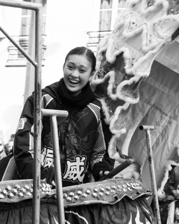 Alice · 01/03/2026 · 3586 x 4482 Cette photographie en noir et blanc met en scène une femme souriante, sujet principal, vêtue d’une tenue traditionnelle chinoise. Elle est positionnée derrière une structure métallique, probablement un décor de théâtre ou de cinéma.Le cadrage est serré sur son visage et sa partie supérieure du corps, avec un arrière-plan flou et des éléments décoratifs stylisés. La lumière semble douce et naturelle, mettant en valeur son expression joyeuse. Les tons dominants sont les gris et noirs, typiques du noir et blanc, renforçant une ambiance rétro ou historique. Le style évoque un contexte théâtral ou cinématographique des années passées.