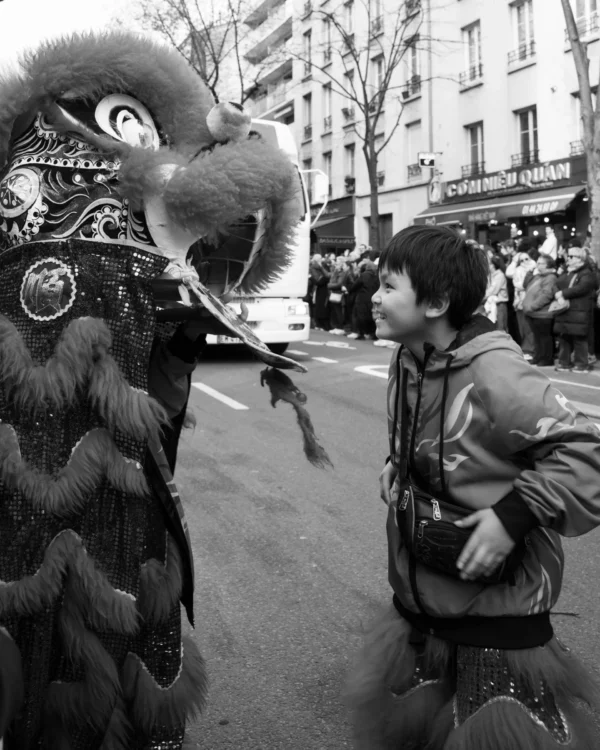 Alice · 01/03/2026 · 3803 x 4754 Cette photographie en noir et blanc capture une scène de rue animée. Le **sujet principal** est un masque ou costume de lion/dragon, imposant et détaillé, face à une personne en veste qui semble interagir avec lui. La **composition** est dynamique, avec un cadrage serré sur les visages et les détails des costumes. La **lumière** est naturelle, contrastée, mettant en valeur les textures et les expressions. Les **couleurs dominantes** sont absentes en raison du filtre noir et blanc, mais les contrastes renforcent les ambiances. Le **style** est documentaire et spontané, évoquant une **ambiance** festive et culturelle, probablement liée à une célébration traditionnelle.