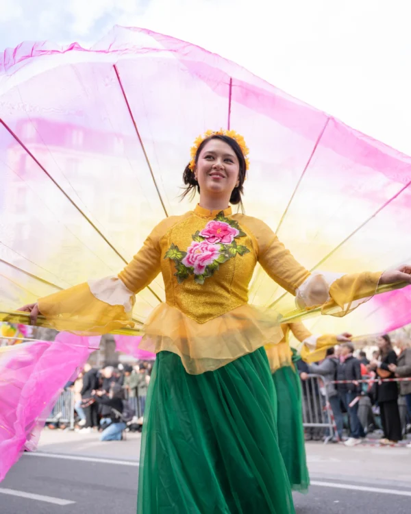 Alice · 01/03/2026 · 4314 x 5392 Cette photographie met en scène une femme comme sujet principal, vêtue d’une tenue colorée (robe verte et jaune) et tenant de grands éventails aux teintes pastel (rose et jaune). La composition est dynamique, avec un cadrage serré sur elle, en mouvement.La lumière est naturelle et douce, mettant en valeur les couleurs vives. Les tons dominants sont les verts, jaunes et roses, créant une ambiance festive et joyeuse. Le style évoque une célébration culturelle ou un défilé, renforcé par l’arrière-plan flou et les spectateurs en arrière. L’ambiance est joyeuse et énergique.