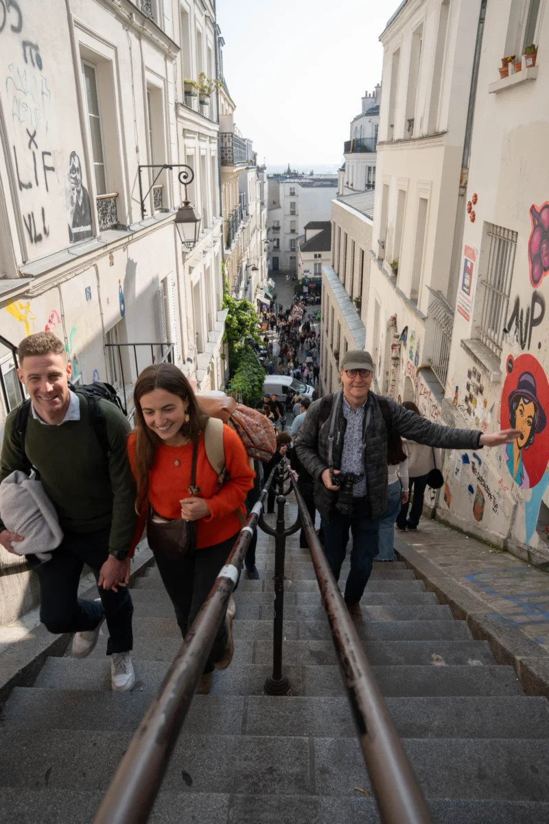 Alice · 21/03/2026 · 4672 x 7008 Cette photographie montre quatre personnes marchant sur un escalier métallique en colimaçon dans une rue étroite et pentue, probablement à Paris. Le sujet principal est le groupe, souriant et détendu.La composition utilise un cadrage serré sur les marches, avec un arrière-plan de bâtiments anciens et de graffitis. La lumière est naturelle, douce, mais légèrement voilée, typique d’un jour nuageux. Les couleurs dominantes sont les tons neutres des bâtiments, contrastés par les vêtements colorés des individus (orange, noir, gris).L’ambiance est conviviale et dynamique, évoquant une balade touristique dans un quartier pittoresque.