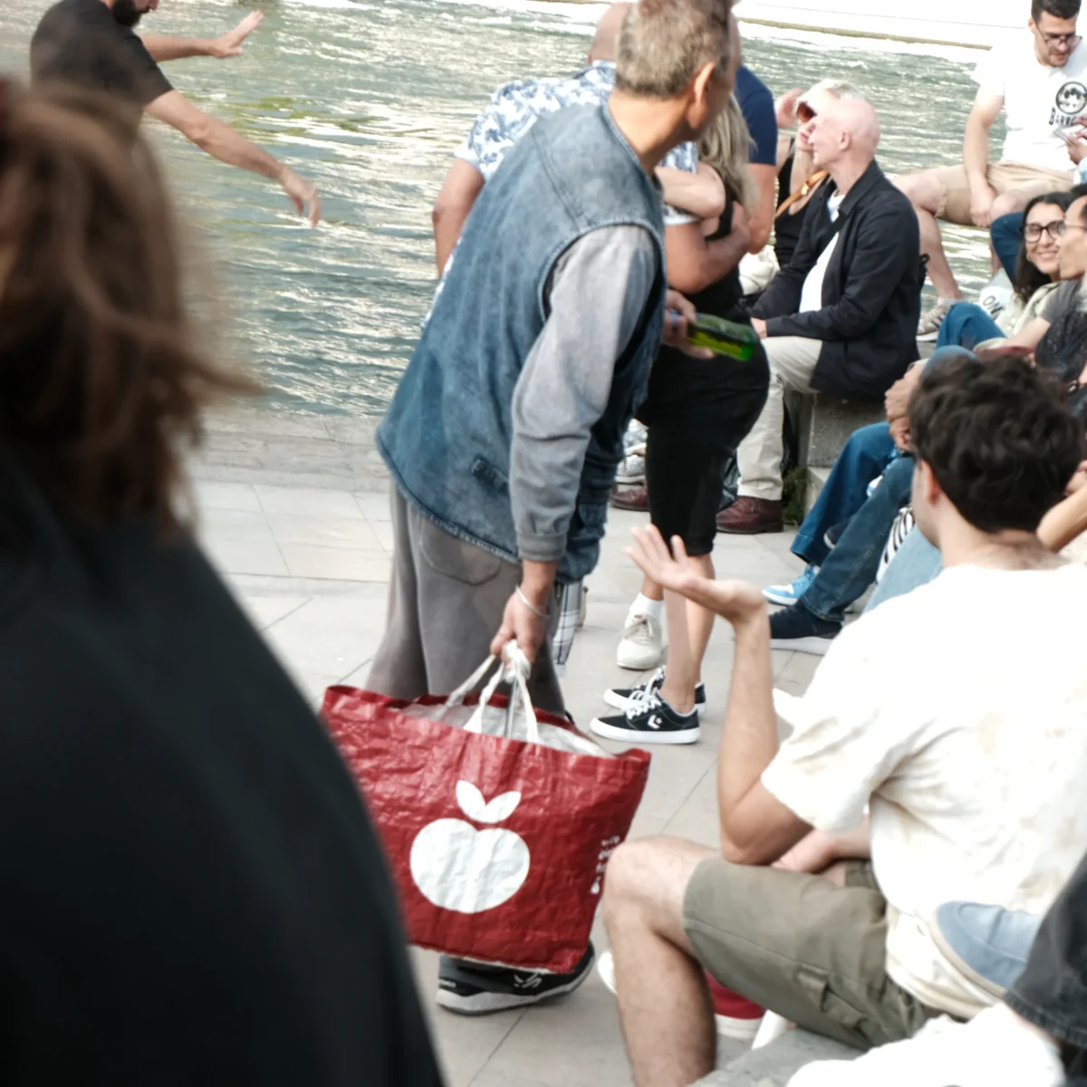 Nathalie F. · 02/08/2025 · 4160 x 4160 Cette photographie montre un groupe de personnes en bordure d’eau, probablement un quai ou un bord de rivière. Le sujet principal semble être un homme en jean et veste grise, entouré d’autres individus assis ou debout.La composition est dynamique, avec un cadrage serré sur le premier plan où un sac rouge à motifs blancs est visible. L’ambiance est informelle et détendue. La lumière semble naturelle, légèrement diffuse, créant des ombres douces. Les couleurs dominantes sont les tons neutres (gris, beige) et quelques touches de rouge et bleu. Le style est documentaire, capturant un moment du quotidien.