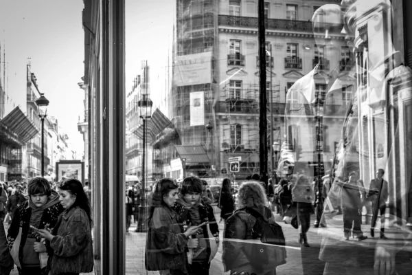 AnJo Beckers · 07/03/2026 · 5382 x 3588 Cette photographie en noir et blanc capture une rue animée vue à travers une vitrine. Le sujet principal est la foule de passants, habillés chaudement, suggérant un climat froid. La composition montre une perspective symétrique avec des reflets et des ombres créant une ambiance dynamique. Le cadrage serré met en valeur les détails des visages et des mouvements.La lumière est douce, avec des contrastes marqués entre ombres et clartés. Les couleurs dominantes, bien que monochromes, jouent sur les nuances de gris. Le style est documentaire et réaliste, évoquant une ambiance urbaine et vivante, presque cinématographique.