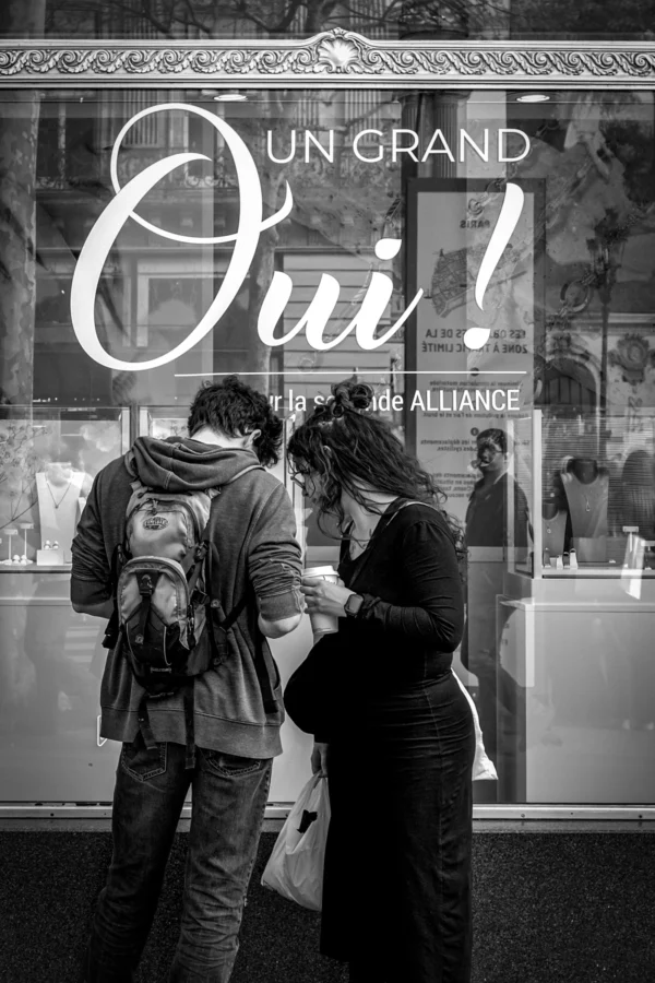 AnJo Beckers · 07/03/2026 · 2989 x 4483 Cette photographie en noir et blanc montre deux personnes discutant devant une vitrine. Le sujet principal est l'échange entre elles, accentué par leur proximité et leur posture tournée l'une vers l'autre. La composition utilise un cadrage serré sur les deux individus, avec la vitrine en arrière-plan portant l'inscription "Un grand Oui!" en lettres stylisées.La lumière douce et contrastée crée des ombres marquées, renforçant l'ambiance intimiste. Les tons dominants sont les gris et noirs, typiques du monochrome, ajoutant une touche dramatique et élégante. Le style est sobre et photographique, évoquant une scène de vie quotidienne avec une ambiance à la fois moderne et intemporelle.