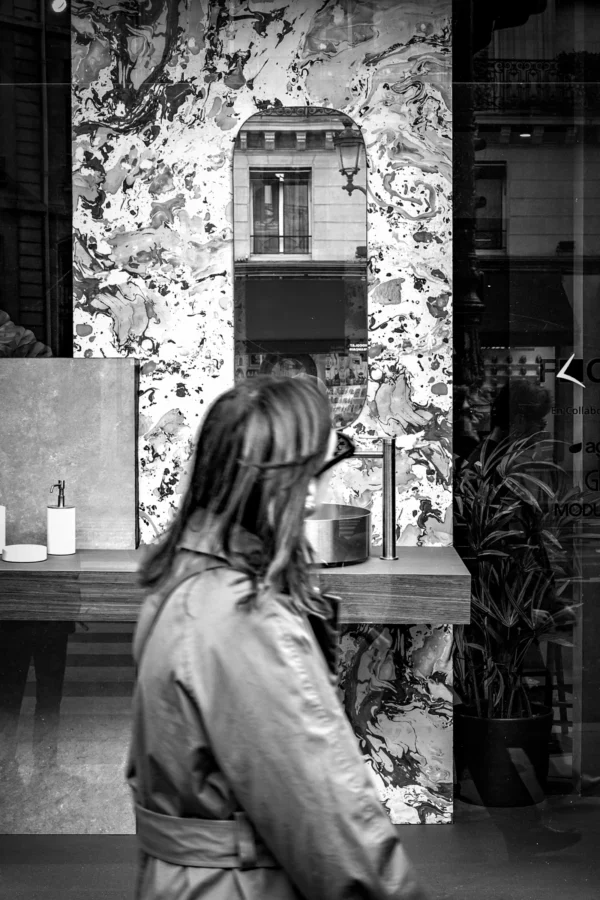 AnJo Beckers · 07/03/2026 · 3139 x 4708 Cette photographie en noir et blanc met en scène une personne de dos, penchée sur un comptoir, semblant observer ou manipuler des objets. Le sujet principal est flou, attirant l’attention vers l’arrière-plan où un petit bâtiment mural et des éléments décoratifs sont visibles.La composition joue sur les contrastes de profondeur et de netteté. Le cadrage est serré, centré sur la silhouette et l’espace immédiat. La lumière, douce et tamisée, crée des ombres douces et une ambiance mystérieuse. Le style, sobre et artistique, évoque une touche documentaire ou conceptuelle. L’ambiance est intime et contemplative.