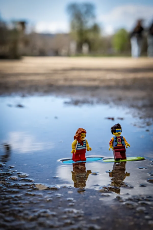AnJo Beckers · 14/03/2026 · 4000 x 6000 Cette photographie met en scène deux figurines Lego habillées en surfeurs, posées sur une surface humide reflétant leur image. Le sujet principal est ces deux personnages, placés au centre de la composition.Le cadrage est serré, mettant l’accent sur les figurines. La lumière naturelle, douce et légèrement dorée, crée des ombres discrètes. Les couleurs dominantes sont les tons froids (bleu, gris) et les couleurs vives des Lego (jaune, rouge, vert).Le style est minimaliste et ludique, évoquant une ambiance à la fois poétique et humoristique.