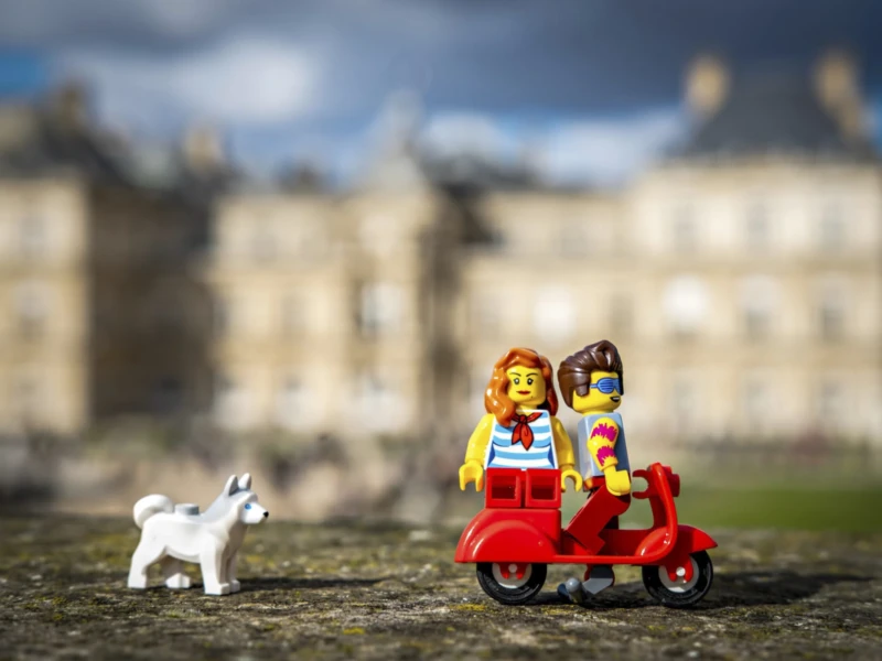 Mondes minuscules au jardin du Luxembourg Cette photographie présente un diorama miniature mettant en scène deux figurines Lego, un personnage masculin et un autre féminin, assis sur un véhicule rouge. À gauche, un petit chien blanc Lego les accompagne. Le cadrage est serré sur ces éléments, avec en arrière-plan un bâtiment historique flou, probablement un château.La lumière est naturelle, douce et légèrement contrastée, créant des ombres douces. Les couleurs dominantes sont les tons rouges du véhicule, les teintes pastel des figurines et les gris/beiges du décor. Le style est ludique et artistique, évoquant une ambiance joyeuse et nostalgique.