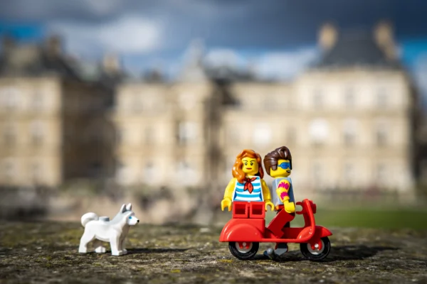AnJo Beckers · 14/03/2026 · 5574 x 3716 Cette photographie présente un sujet principal composé de deux figurines Lego, un garçon et une fille, assis dans un petit véhicule rouge, accompagnés d'un chien blanc en Lego également. La composition est simple et centrée, avec un cadrage serré sur les figurines.L'arrière-plan flou montre un bâtiment architectural classique, suggérant un cadre historique. La lumière est naturelle, douce et légèrement contrastée, mettant en valeur les couleurs vives des figurines. Les couleurs dominantes sont le rouge du véhicule, les tons pastel des vêtements des figurines et le blanc du chien. Le style est ludique et miniature, créant une ambiance joyeuse et fantaisiste.