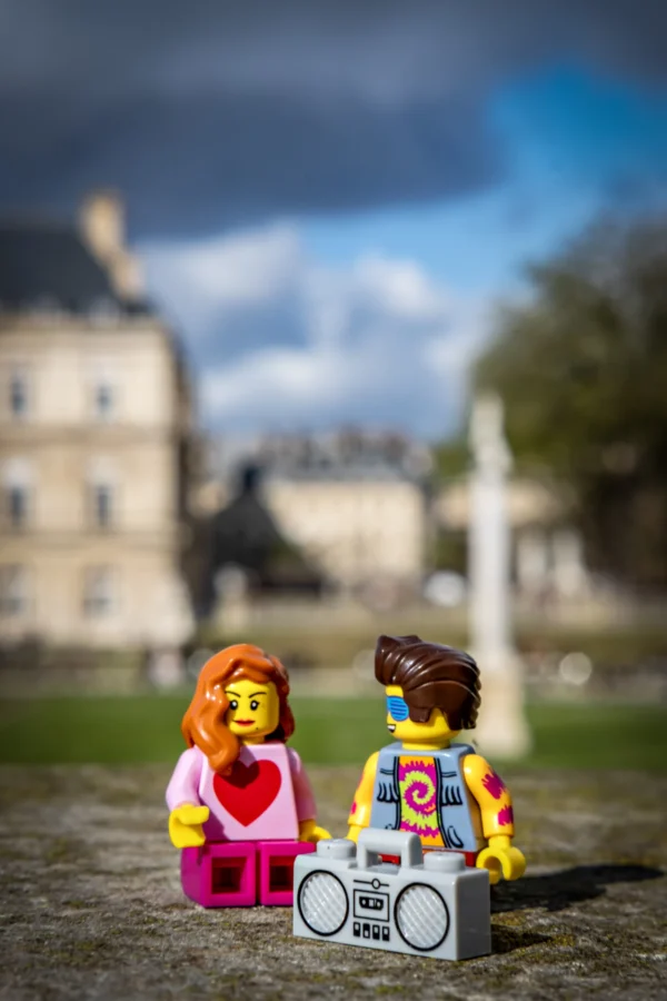 AnJo Beckers · 14/03/2026 · 3497 x 5246 Cette photographie met en scène deux figurines LEGO en premier plan, représentant deux personnages stylisés : une femme en robe rose et un homme avec un boombox. La composition joue sur le contraste entre ces figurines miniatures et l'arrière-plan flou d'un bâtiment classique et d'un parc.Le cadrage est serré sur les figurines, avec une profondeur de champ réduite. La lumière est naturelle, douce et légèrement contrastée, mettant en valeur les couleurs vives des LEGO. Les couleurs dominantes sont les tons pastel et primaires des figurines, créant une ambiance ludique et joyeuse malgré le cadre architectural sérieux. Le style est à la fois minimaliste et créatif.