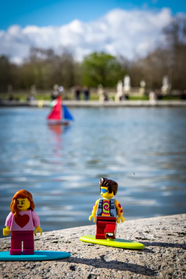 AnJo Beckers · 14/03/2026 · 3296 x 4944 Cette photographie met en scène deux figurines LEGO sur des planches de surf, posées sur un bord de lac. Le sujet principal est ces deux personnages miniatures, l’un en tenue rose, l’autre en maillot coloré.La composition joue sur le contraste entre le premier plan (figurines) et l’arrière-plan flou (personnes nageant). Le cadrage est serré sur les figurines, avec une profondeur de champ réduite.Les couleurs dominantes sont vives (rose, bleu, vert), sous une lumière naturelle et ensoleillée. Le style est ludique et créatif, évoquant une ambiance joyeuse et estivale.