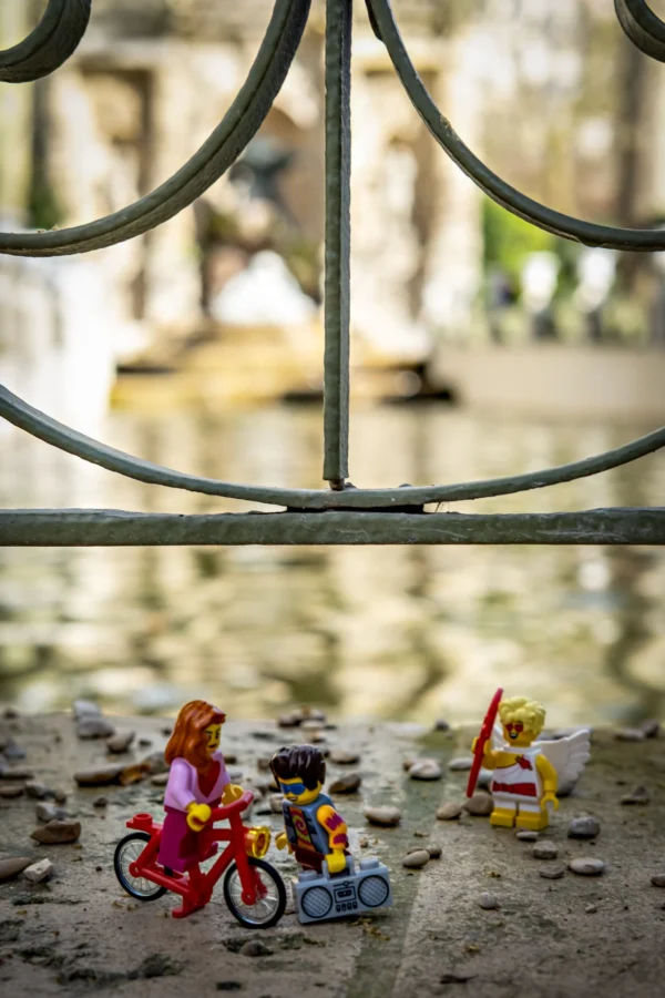 AnJo Beckers · 14/03/2026 · 3221 x 4831 Cette photographie met en scène un **sujet principal** composé de petites figurines Lego représentant des personnages sur des vélos et un tracteur. La **composition** joue sur un **cadrage serré** en bas, contrastant avec un arrière-plan flou et surélevé, montrant une grille métallique et un ciel.La **lumière** est naturelle, douce et légèrement tamisée, créant des ombres douces. Les **couleurs dominantes** sont vives et contrastées : rouge, bleu, jaune et vert, typiques des Lego.Le **style** est épuré et ludique, évoquant une scène miniature. L’**ambiance** est joyeuse et nostalgique, mêlant innocence et créativité.