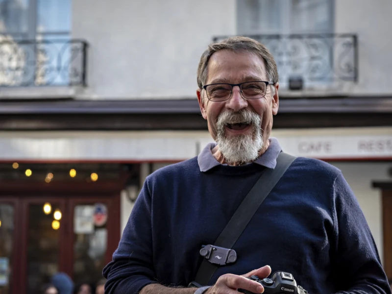Claude, animateur de Portraits de rue Cette photographie met en scène un homme d'âge mûr comme sujet principal, souriant et portant des lunettes, un pull bleu et un appareil photo autour du cou. Le cadrage est serré, centré sur son visage et sa poitrine.La composition est simple et naturelle, avec un arrière-plan flou montrant une façade de bâtiment et des enseignes. La lumière est douce et naturelle, probablement en journée, éclairant le sujet de face. Les couleurs dominantes sont les tons bleus du pull et les teintes neutres de l'arrière-plan. Le style est réaliste et spontané, créant une ambiance chaleureuse et conviviale.