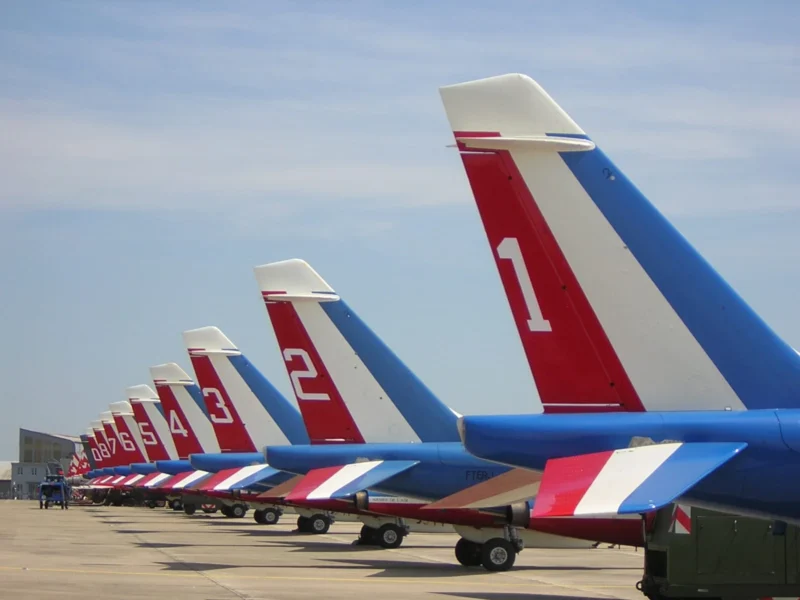 Frédéric · 11/06/2006 · 1600 x 1200 Cette photographie met en scène une rangée d’avions de chasse alignés. Le **sujet principal** est leur queue colorée, avec des motifs **bleus, blancs et rouges** distinctifs.La **composition** est symétrique et linéaire, renforçant l’harmonie visuelle. Le **cadrage** serré sur les avions crée une perspective dynamique. La **lumière** est naturelle, probablement en plein jour, avec un éclairage uniforme.Les **couleurs dominantes** sont vives et contrastées, évoquant une ambiance **militaire et professionnelle**. Le style est **documentaire et technique**, transmettant une impression d’ordre et de précision. L’ambiance est à la fois **puissante et disciplinée**.