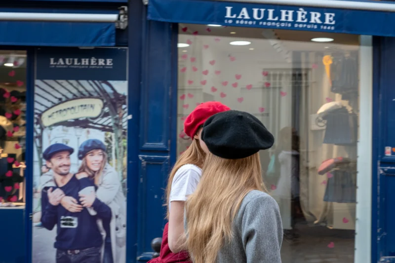 Jean-Marc · 21/03/2026 · 5281 x 3521 Cette photographie montre deux femmes devant une boutique **Laulhère**. La personne au premier plan, dos à la caméra, porte un béret rouge et une veste grise. La composition met en valeur l’entrée de la boutique, avec des affiches publicitaires et des mannequins habillés en uniformes.Le cadrage est serré, centré sur les deux femmes et l’entrée du magasin. La lumière est naturelle et douce, sans ombres marquées. Les couleurs dominantes sont les tons bleus (boutique), gris (vêtements), et rouge (béret).L’ambiance est urbaine et élégante, évoquant un style parisien classique et moderne.