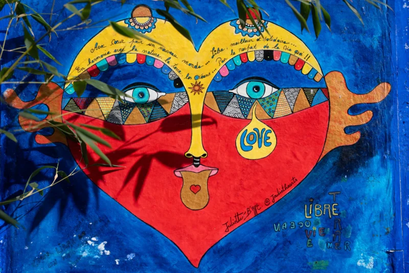 Jean-Marc · 21/03/2026 · 6048 x 4032 Cette photographie met en scène un cœur coloré et stylisé, sujet principal, peint sur un support mural bleu. Le cœur est décoré de motifs variés : yeux géométriques, mains et éléments graphiques colorés. La composition est symétrique, avec des éléments artistiques et symboliques.Le cadrage est serré, centré sur le cœur. La lumière est naturelle, mettant en valeur les couleurs vives et les détails. Les couleurs dominantes sont le bleu, le rouge, le jaune et le vert, créant un style artistique et joyeux. L’ambiance est festive, positive et pleine d’énergie.