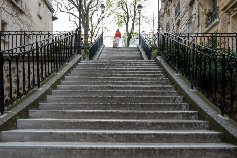 Jean-Marc · 21/03/2026 · 5357 x 3571 Cette photographie montre une personne en haut d’un escalier extérieur en pierre, vêtue d’un manteau rouge. La composition met en valeur les marches descendantes, bordées de rampes métalliques noires. Le cadrage est serré sur les escaliers, créant une perspective profonde.La lumière est naturelle, légèrement tamisée, évoquant un jour d’hiver. Les couleurs dominantes sont les tons gris (pierres, ciel) et noir (rampes), rehaussés par le rouge vif du manteau. Le style est épuré et réaliste, avec une ambiance calme et légèrement mélancolique, renforcée par l’absence de mouvement et la froideur des matériaux.