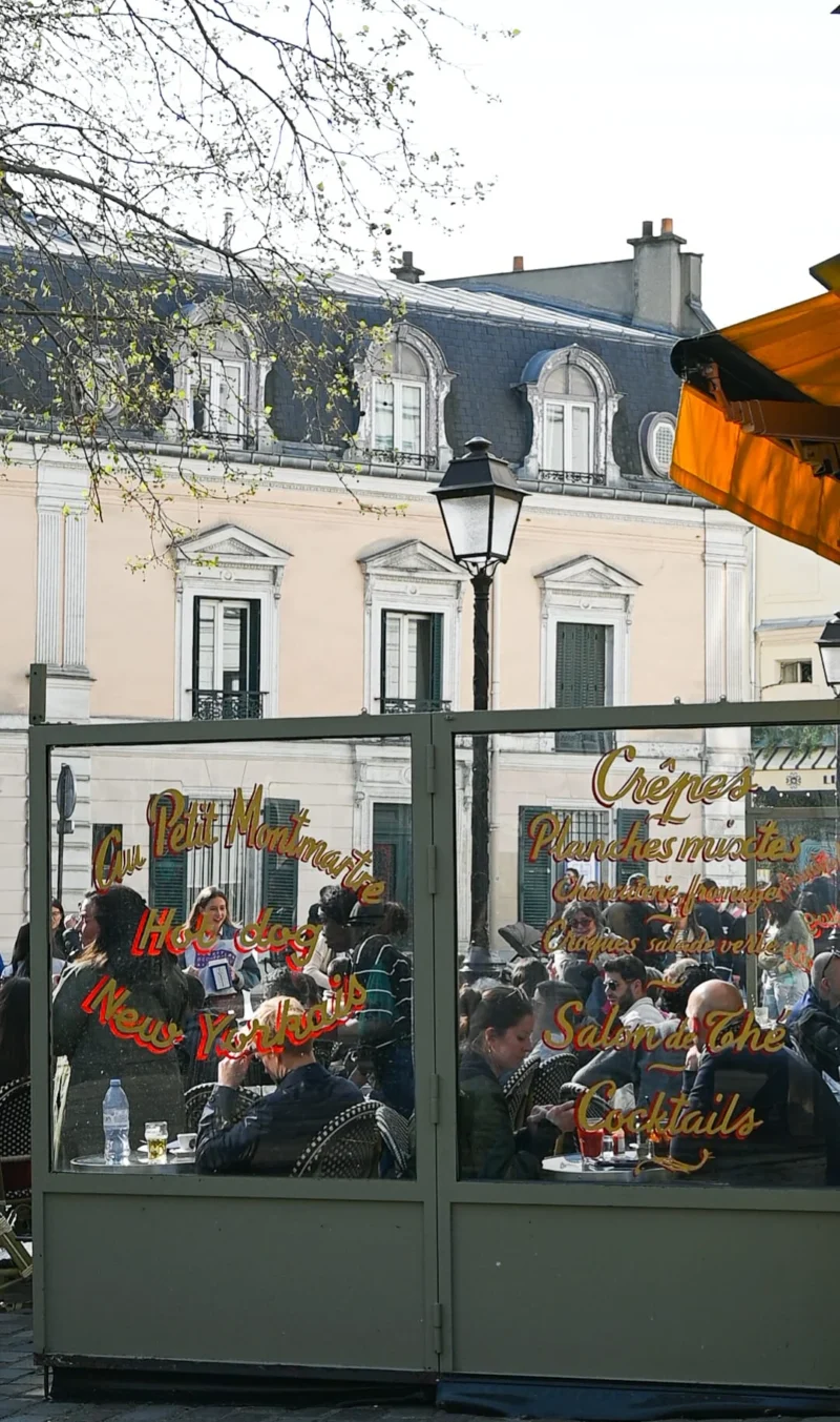Aurélie · 01/01/2021 · 1080 x 1827 Cette photographie capture une scène de terrasse de café en extérieur. Le sujet principal est un groupe de personnes assises sous des parasols orange, discutant et profitant de leur boisson.La composition montre une vue en profondeur, avec un cadrage serré sur les clients et un arrière-plan de bâtiments classiques. La lumière est naturelle et douce, typique d'une journée ensoleillée. Les couleurs dominantes sont les tons chauds (orange des parasols, beige des murs) et les verts des plantes grimpantes.Le style est réaliste et spontané, évoquant une ambiance conviviale et détendue, caractéristique des cafés urbains.