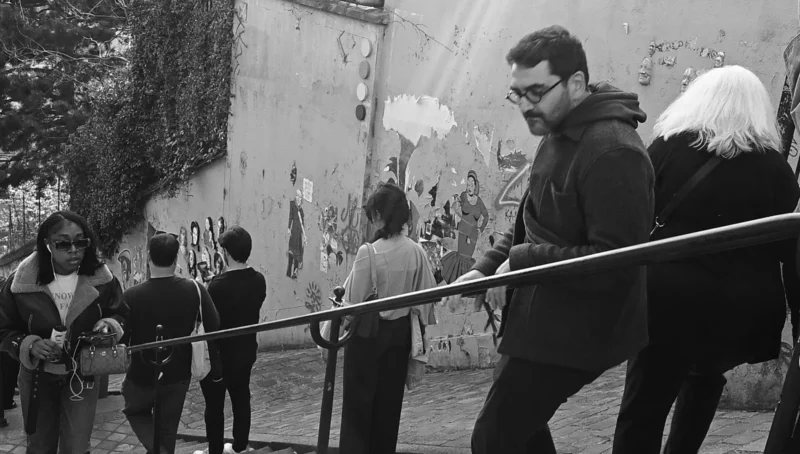 Aurélie · 01/01/2021 · 1902 x 1080 Cette photographie en noir et blanc montre un homme comme sujet principal, marchant devant un mur tagué. La composition inclut plusieurs personnes en arrière-plan, dont certaines semblent observer ou discuter.Le cadrage est serré sur l’homme, avec une profondeur de champ qui met en valeur les détails du mur et les silhouettes. La lumière est naturelle, douce, sans contraste marqué, créant une ambiance sobre et réaliste.Les couleurs dominantes sont les tons de gris, typiques du noir et blanc. Le style est documentaire, captant une scène urbaine spontanée. L’ambiance est calme et légèrement mélancolique, reflétant un moment de vie quotidienne dans un environnement urbain.