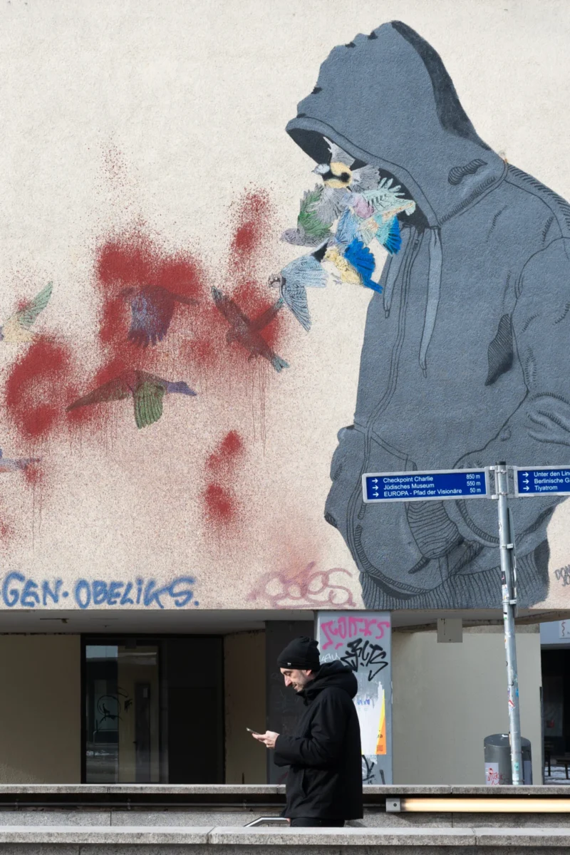 Daniel · 20/02/2026 · 2035 x 3052 Cette photographie montre un graffiti mural dominant le cadre, représentant une silhouette en hoodie tenant un masque à gaz, avec des éclaboussures rouges évoquant du sang. En bas, une personne marche devant un bâtiment aux couleurs neutres, avec une enseigne "GEN-OBELIX".La composition oppose le graffiti sombre et expressif à la scène urbaine plus discrète. Le cadrage est serré sur le mural, avec une lumière naturelle diffuse. Les couleurs dominantes sont les tons gris, bleu et rouge sang. Le style est street art réaliste, créant une ambiance à la fois inquiétante et engagée, reflétant une critique sociale ou politique.