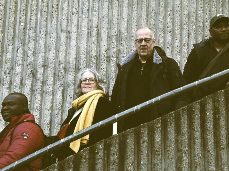 Terry, AnJo, Franck et Don de Photos in Paris Cette photographie montre quatre personnes sur un escalier métallique extérieur. Le sujet principal semble être l’homme au centre, vêtu d’une robe noire, probablement un religieux. La composition est dynamique, avec un cadrage serré sur les visages et les épaules.La lumière est naturelle, légèrement tamisée, créant des contrastes doux. Les couleurs dominantes sont les tons froids (gris du métal et du mur) et les couleurs chaudes (vêtements rouges et jaune). Le style est documentaire, réaliste, avec une ambiance sobre et contemplative, évoquant une scène de vie quotidienne ou un moment solennel.