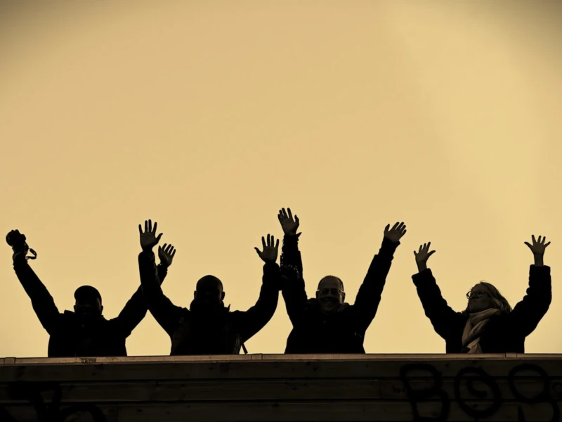 Photos in Paris Cette photographie met en scène un groupe de personnes en silhouette, bras levés, comme pour applaudir ou célébrer. Le sujet principal est leur silhouette unie, créant une impression de solidarité.La composition est symétrique et horizontale, avec les personnages alignés sur une balustrade. Le cadrage est serré, centré sur eux.La lumière douce et dorée, probablement un coucher de soleil, donne une ambiance chaleureuse et dramatique. Les couleurs dominantes sont les tons chauds de jaune et orange, renforçant l’émotion.Le style est épuré et cinématographique, évoquant une ambiance à la fois joyeuse et intense.