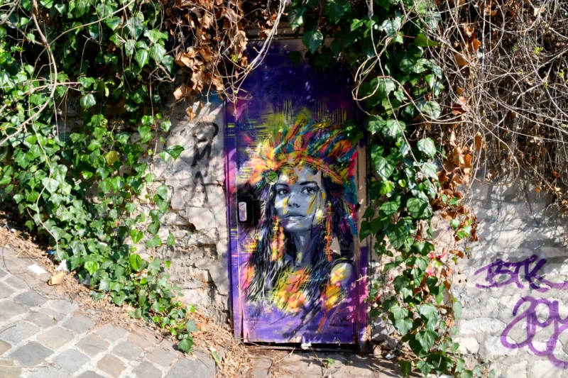 Vincent · 21/03/2026 · 5568 x 3712 Cette photographie montre une boîte aux lettres ou un abri mural recouvert de graffitis colorés et de tags. Le sujet principal est cette surface taguée, entourée de végétation luxuriante.La composition est simple, avec un cadrage serré mettant en valeur les couleurs vives et les motifs artistiques. La lumière naturelle éclaire la scène, accentuant les contrastes entre les tons vifs des graffitis et le vert des plantes. Les couleurs dominantes sont les violets, jaunes, verts et rouges, créant un style urbain et dynamique. L’ambiance est à la fois artistique et sauvage, reflétant une touche de créativité dans un environnement naturel.