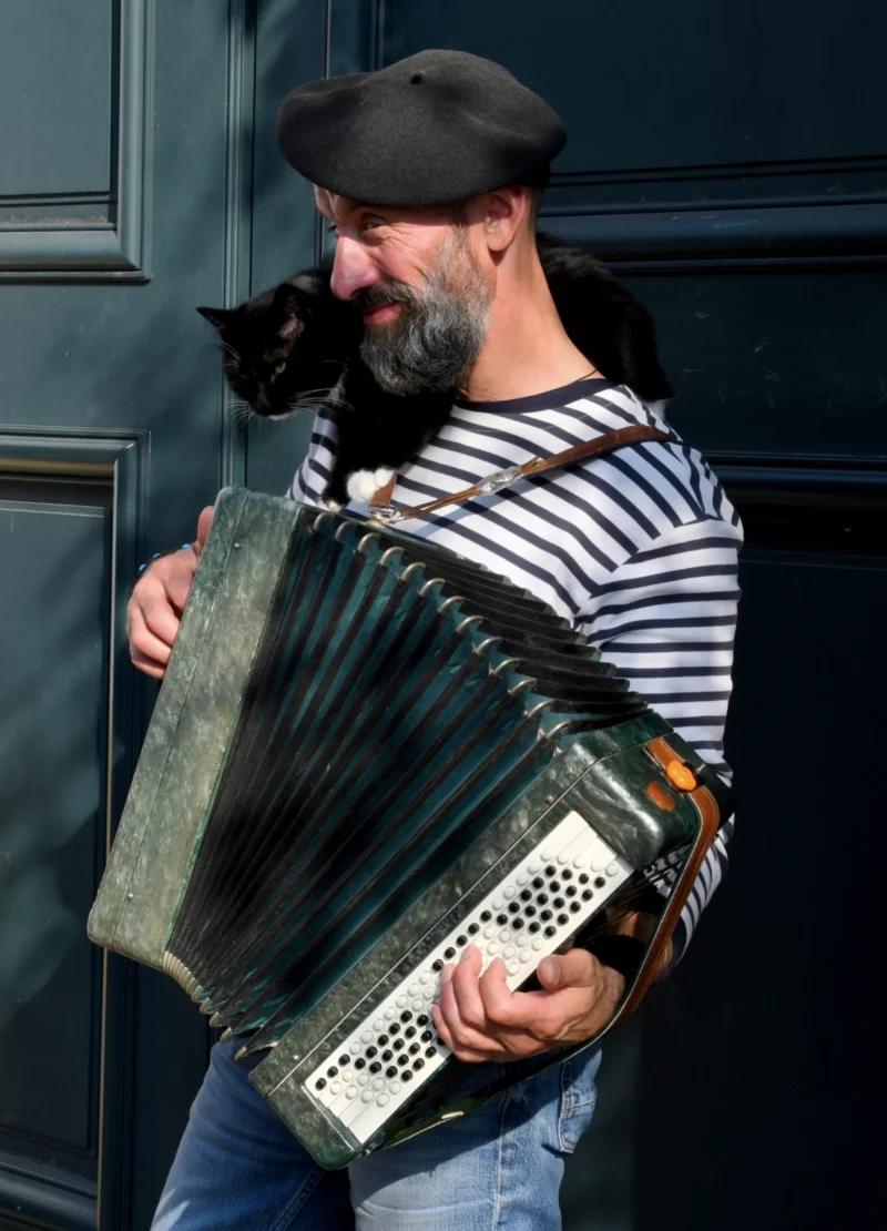 Vincent · 21/03/2026 · 3319 x 4605 Cette photographie met en scène un homme comme sujet principal, jouant de l'accordéon. Il porte un béret, un pull à rayures horizontales et une barbe. Un chat noir est posé sur son épaule.La composition est naturelle et spontanée, avec un cadrage serré sur le haut du corps. La lumière semble douce et naturelle, éclairant le sujet de manière homogène. Les couleurs dominantes sont les tons neutres (gris, noir) et les rayures blanches et bleues du pull.Le style est réaliste et décontracté, créant une ambiance chaleureuse et intimiste.