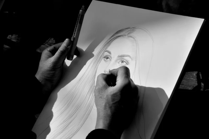 Vincent · 21/03/2026 · 5568 x 3712 Cette photographie montre une personne en train de dessiner un portrait au fusain ou à la craie sur une feuille fixée sur une planche. Le **sujet principal** est le geste artistique en cours.La **composition** est serrée, centrée sur les mains et le visage esquissé. Le **cadrage** est rapproché, mettant l’accent sur le processus créatif. La **lumière** est contrastée, avec des ombres marquées, renforçant l’aspect dramatique. Les **couleurs dominantes** sont en noir et blanc, accentuant les textures et les contrastes.Le **style** est réaliste et spontané, évoquant une **ambiance** intime et concentrée.