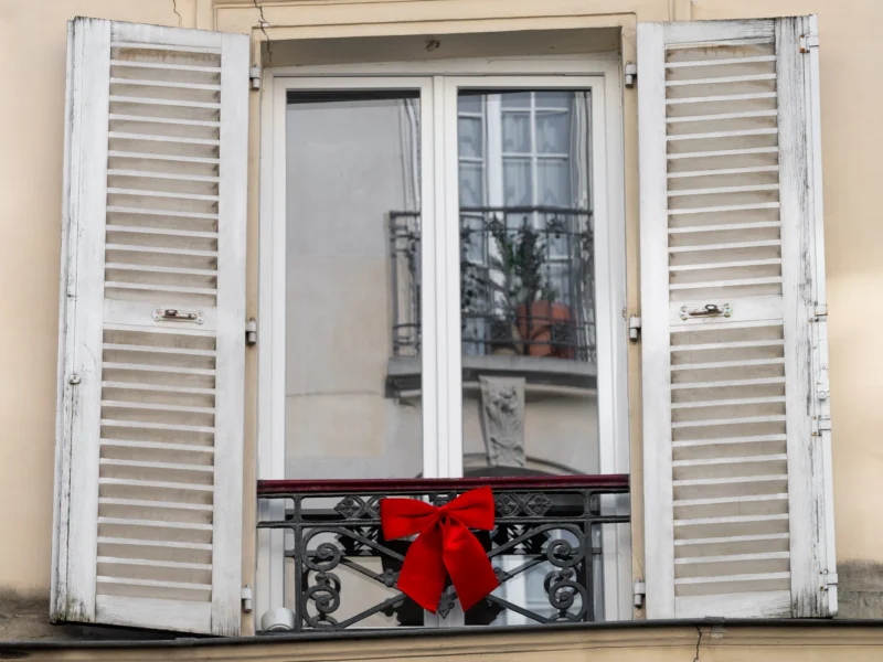 Daniel · 21/03/2026 · 3850 x 2888 Cette photographie montre une fenêtre à volets blancs entrouverts, décorée d’un gros nœud rouge accroché à la balustrade en fer forgé. La composition est simple et centrée, avec un cadrage serré sur la fenêtre.La lumière semble naturelle, douce et légèrement tamisée, créant des ombres subtiles. Les couleurs dominantes sont les tons neutres (blanc des volets, beige du mur) contrastés par le rouge vif du nœud.Le style est épuré et réaliste, évoquant une ambiance chaleureuse et festive, typique d’une décoration de Noël ou d’un événement spécial.