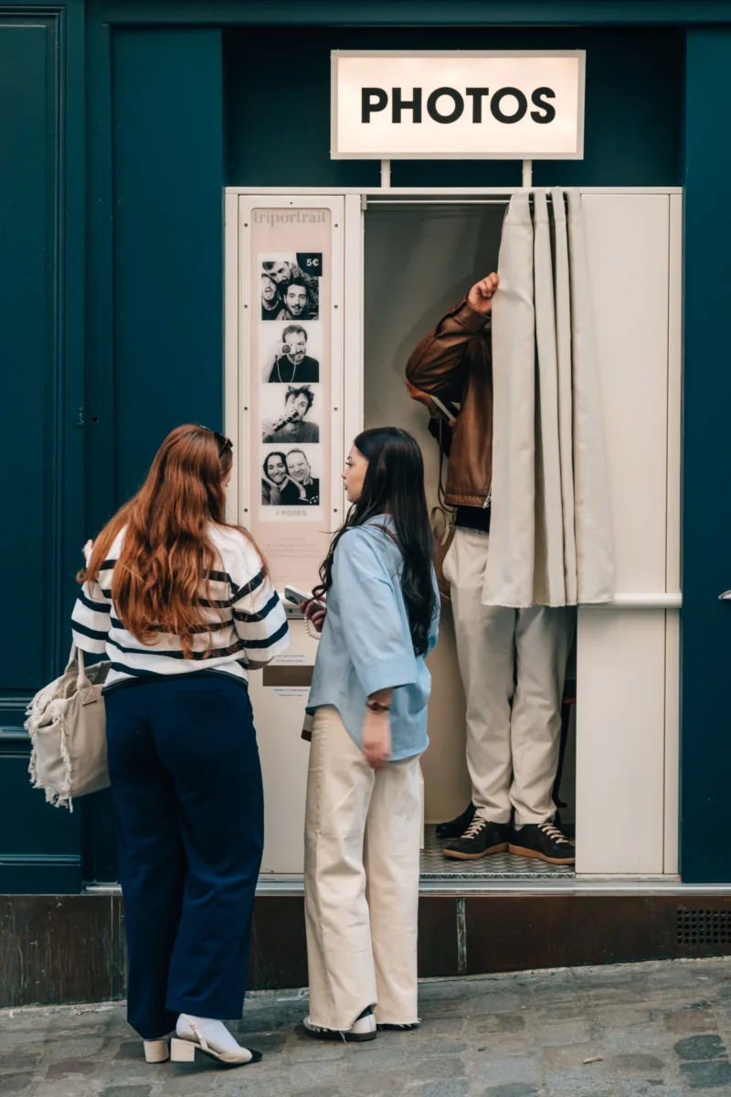 Daniel · 21/03/2026 · 3572 x 5358 Cette photographie montre deux femmes observant des photos exposées dans une vitrine. Le sujet principal est leur interaction avec l’affichage.La composition est symétrique, avec les deux femmes placées côte à côte devant la vitrine. Le cadrage est serré sur elles et l’exposition.La lumière est naturelle et douce, éclairant les visages et les détails des photos. Les couleurs dominantes sont les tons froids (bleu de la façade) et chauds (vêtements des femmes).Le style est réaliste et documentaire, capturant un moment quotidien. L’ambiance est calme et intrigante, évoquant la curiosité et l’échange.