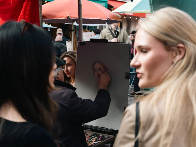 Daniel · 21/03/2026 · 3386 x 2539 Cette photographie montre deux femmes observant une œuvre d’art en cours de création, probablement un portrait. Le sujet principal est le tableau et les mains de l’artiste qui le dessine.La composition est serrée, centrée sur les visages et le travail artistique. Le cadrage est en plan moyen, mettant en valeur les interactions et les détails.La lumière semble naturelle et douce, éclairant les sujets sans ombres marquées. Les couleurs dominantes sont neutres, avec des touches de rouge et de vert en arrière-plan.L’ambiance est conviviale et artistique, évoquant un événement culturel ou une exposition.