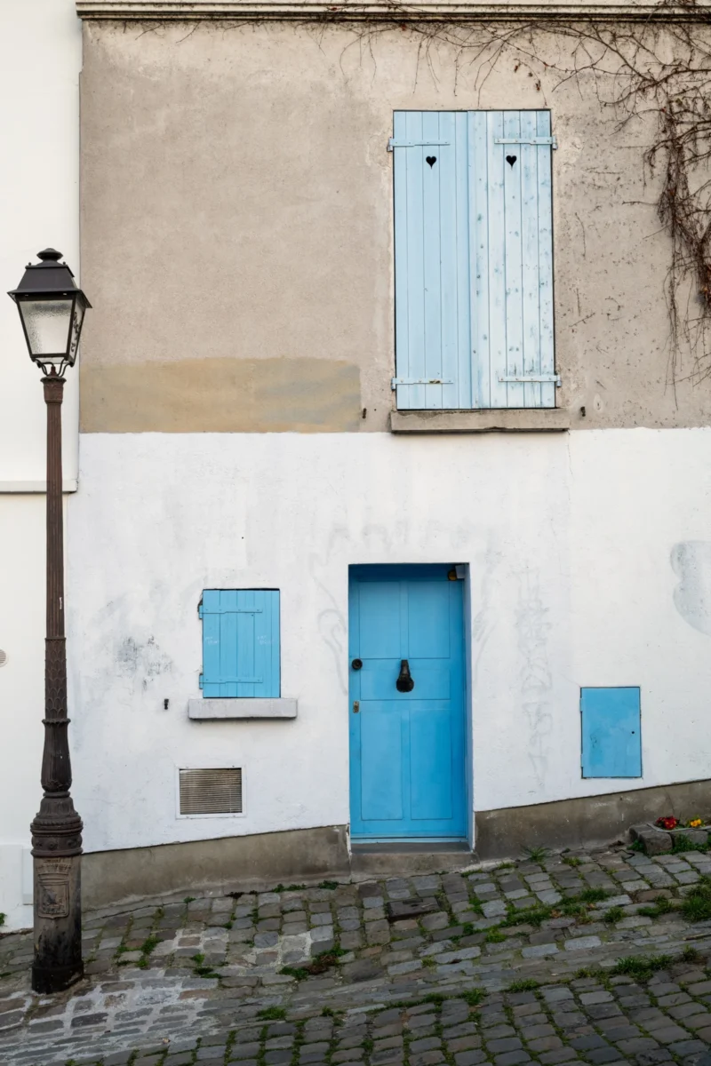 Daniel · 21/03/2026 · 3867 x 5800 Cette photographie montre une façade d’immeuble aux tons pastel. Le sujet principal est une porte bleue et des volets identiques, partiellement ouverts. La composition est simple et symétrique, avec un cadrage serré sur l’entrée.La lumière naturelle, douce, met en valeur les textures et les dégradés des couleurs. Les teintes dominantes sont le bleu clair, le beige et le gris, renforçant une ambiance sobre et apaisante. Le style évoque un charme urbain et vintage, typique des quartiers historiques. L’ambiance est calme et intemporelle, soulignée par les pavés et la lampe ancienne.