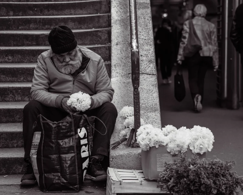 Alice · 21/03/2026 · 4355 x 3484 Cette photographie en noir et blanc met en scène un homme âgé assis sur une marche, le sujet principal. Il semble fatigué, les mains posées sur ses genoux.La composition est simple et centrée, avec un cadrage serré sur le vieil homme et ses fleurs blanches à côté de lui. En arrière-plan, une personne floue et un environnement urbain sont visibles.La lumière est douce, créant des contrastes modérés. Les tons dominants sont les gris et noirs, adoucis par les blancs des fleurs.Le style est documentaire et réaliste, évoquant une ambiance mélancolique et contemplative.
