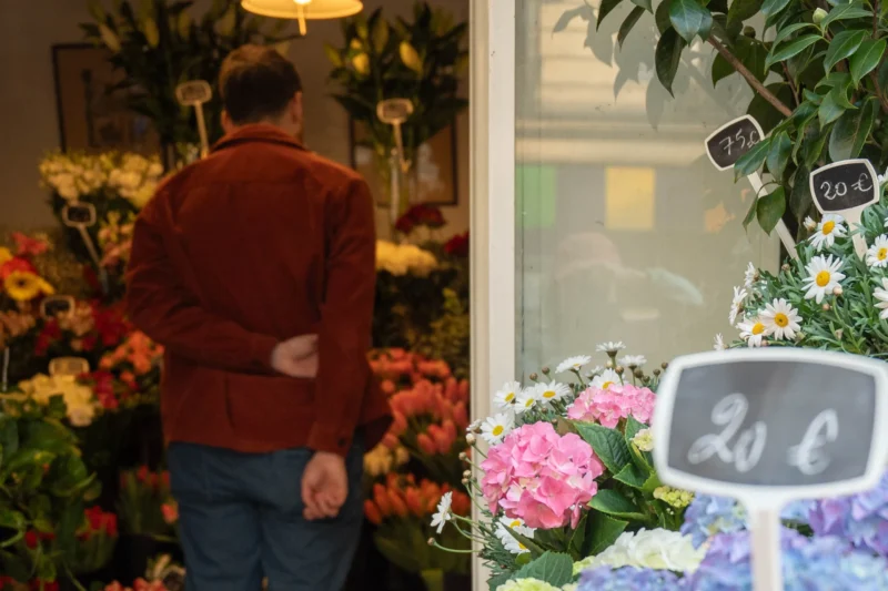 Alice · 21/03/2026 · 2713 x 1809 Cette photographie montre un homme de dos, observant des fleurs dans un magasin. La composition est divisée en deux parties : à gauche, l’homme et les bouquets colorés, à droite, une vitrine avec des fleurs et un panneau affichant un prix (20 €).Le cadrage est serré sur les détails. La lumière est douce et naturelle, mettant en valeur les couleurs vives des fleurs. Les teintes dominantes sont les tons chauds (rouges, roses) et frais (blancs, verts). Le style est réaliste et quotidien, créant une ambiance chaleureuse et accueillante, typique d’un commerce de fleurs.