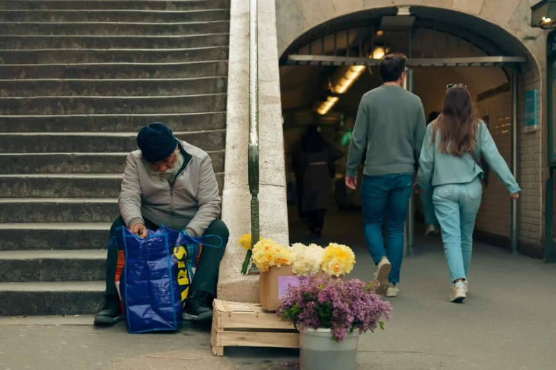 Don · 21/03/2026 · 1536 x 1024 Cette photographie met en scène un homme penché sur un sac plastique bleu, près d’un étalage de fleurs. La composition joue sur les contrastes entre l’avant-plan (l’homme et les fleurs) et l’arrière-plan (les personnes marchant sous une arche).Le cadrage est serré sur le sujet principal, avec une profondeur de champ qui floute légèrement l’arrière-plan. La lumière est naturelle et douce, créant des ombres douces. Les couleurs dominantes sont les tons froids (bleu, gris) et chauds (jaunes, violets des fleurs).L’ambiance est réaliste et quotidienne, évoquant une scène de marché ou de rue. Le style est sobre et documentaire.