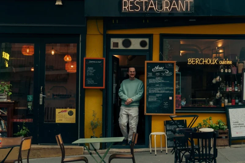 Don · 21/03/2026 · 1536 x 1024 Cette photographie met en scène un homme debout devant un restaurant nommé "Berghof". Le sujet principal est cet homme, vêtu d'un manteau sombre, positionné légèrement en contre-jour.La composition est symétrique avec des tables et des chaises disposées devant le restaurant. Le cadrage est serré, centré sur l'entrée éclairée. La lumière est douce et tamisée, créant une ambiance chaleureuse et nocturne. Les couleurs dominantes sont les tons jaunes et noirs du restaurant, contrastant avec les teintes sombres de la nuit.Le style est réaliste et urbain, évoquant une ambiance calme et intime.