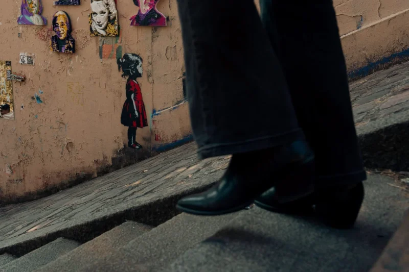 Don · 21/03/2026 · 1536 x 1024 Cette photographie met en scène un **personnage en bas de marches**, partiellement cadré, avec des **pieds et un pantalon visibles**. Le **sujet principal** semble être une fresque murale colorée en arrière-plan, représentant des visages et une silhouette rouge.La **composition** joue sur les contrastes entre l’ombre et la lumière, avec un **cadrage serré** sur les pieds et les escaliers. Les **couleurs dominantes** sont des tons chauds (ocre, rouge) et des contrastes froids (gris des murs et des chaussures).Le **style** est réaliste et urbain, évoquant une **ambiance mystérieuse et artistique**, renforcée par les jeux d’ombres et la présence de graffitis.