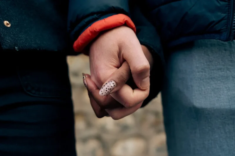 Don · 21/03/2026 · 1536 x 1024 Cette photographie met en scène deux mains entrelacées, l’une portant une bague scintillante. Le sujet principal est l’alliance et le geste intime des mains.La composition est serrée, centrée sur les mains, avec un cadrage rapproché. La lumière est douce et naturelle, créant des ombres subtiles. Les couleurs dominantes sont les tons chauds (rouge du pull, bleu des jeans) et les teintes neutres.Le style est épuré et poétique, évoquant une ambiance tendre et romantique, presque intimiste.