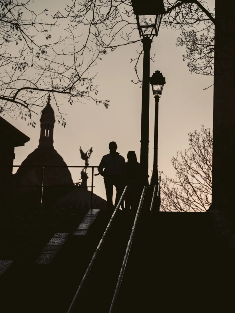 Seila · 21/03/2026 · 3456 x 4608 Cette photographie présente deux silhouettes en contre-jour descendant un escalier. Le sujet principal est ces deux figures, soulignées par leur ombre nette.La composition repose sur une symétrie verticale, avec les lampadaires et les arbres encadrant la scène. Le cadrage est serré, mettant l’accent sur les silhouettes et l’architecture en arrière-plan.La lumière douce et tamisée crée un effet dramatique, typique des clichés en noir et blanc. Les couleurs dominantes sont les tons sépia et gris, renforçant l’ambiance nostalgique et mélancolique. Le style est épuré et poétique, évoquant une atmosphère paisible et intemporelle.