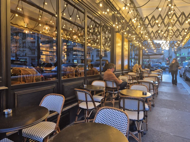Babao · 25/03/2026 · 4000 x 3000 Cette photographie montre un café urbain extérieur. Le sujet principal est l’espace de restauration avec ses tables et chaises disposées sous une structure lumineuse en treillis métallique.La composition est symétrique, avec des tables alignées et des clients assis. Le cadrage inclut une partie de la rue et des vitrines éclairées. La lumière est douce et chaude, provenant des guirlandes lumineuses et des éclairages intérieurs.Les couleurs dominantes sont les tons neutres (noir, gris, blanc) avec des touches de bleu et de bois naturel. L’ambiance est chaleureuse et conviviale, typique d’un café moderne et animé en soirée.
