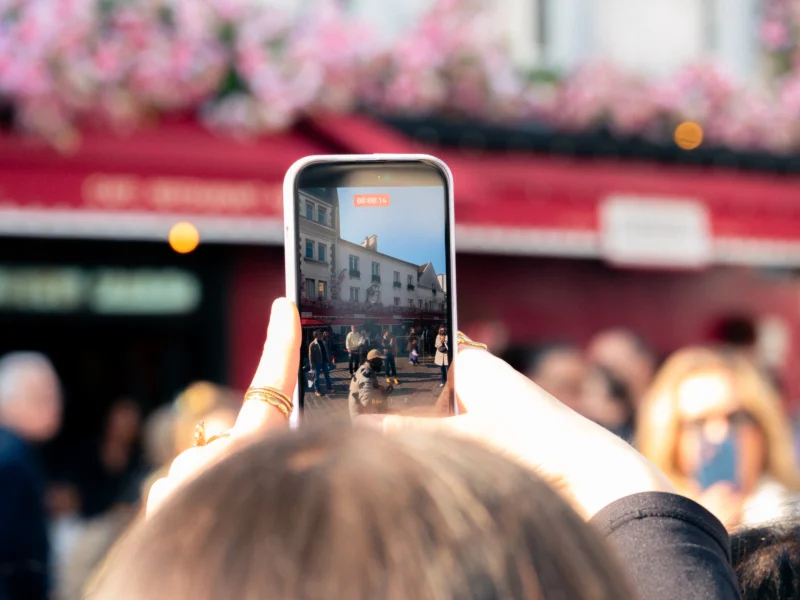 Seila · 21/03/2026 · 4608 x 3456 Cette photographie montre une personne prenant une photo avec son smartphone. Le **sujet principal** est le reflet de l’écran du téléphone, capturant un lieu animé avec des bâtiments et des fleurs roses en arrière-plan.La **composition** est centrée sur le téléphone, avec un **cadrage serré** sur la main et l’écran. La **lumière** est naturelle et éclatante, créant des contrastes marqués. Les **couleurs dominantes** sont les tons chauds (rouge, rose) et les teintes neutres.Le **style** est documentaire et spontané, évoquant une scène du quotidien. L’**ambiance** est dynamique et joyeuse, renforcée par la foule et les fleurs colorées.