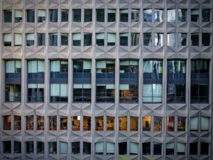 Cyrille · 28/03/2026 · 2026 x 1520 Cette photographie met en scène la façade d’un immeuble moderne aux lignes géométriques marquées. Le sujet principal est l’architecture, avec ses motifs triangulaires en relief et ses fenêtres alignées.La composition repose sur des répétitions symétriques et un cadrage serré, accentuant la structure. La lumière, naturelle et tamisée, crée des contrastes doux entre ombres et reflets sur les vitres. Les couleurs dominantes sont les gris et les tons froids, avec des touches de bleu et de vert reflétées.Le style architectural est contemporain et épuré, évoquant une ambiance à la fois froide et sophistiquée, presque futuriste.