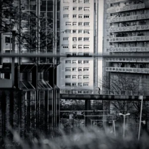 Cyrille · 28/03/2026 · 2026 x 2026 Cette photographie met en scène un ensemble d’immeubles résidentiels modernes en arrière-plan. La composition joue sur les lignes verticales et horizontales, créant une structure géométrique rigoureuse. Le cadrage est serré, avec des éléments architecturaux en premier plan partiellement masqués par des branches.La lumière est tamisée, donnant une ambiance sobre et contrastée. Les tons dominants sont les gris et noirs, renforçant l’aspect monochrome et dramatique. Le style, épuré et minimaliste, évoque une esthétique urbaine contemporaine. L’ambiance est à la fois froide et contemplative, presque mélancolique.