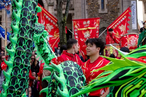 Cyrille · 2026 x 1351 Cette photographie capture une scène festive avec des participants déguisés en dragons ou créatures mythiques. Le sujet principal est un homme souriant, vêtu d’un costume rouge et vert orné d’écailles.La composition montre un groupe en mouvement, avec des éléments colorés comme des bannières rouges et des structures vertes. Le cadrage est serré, mettant en valeur les détails des costumes et les expressions des participants. La lumière semble naturelle, bien répartie, créant des contrastes doux.Les couleurs dominantes sont le rouge et le vert, dynamiques et festives. Le style évoque une célébration traditionnelle ou un carnaval. L’ambiance est joyeuse et énergique.