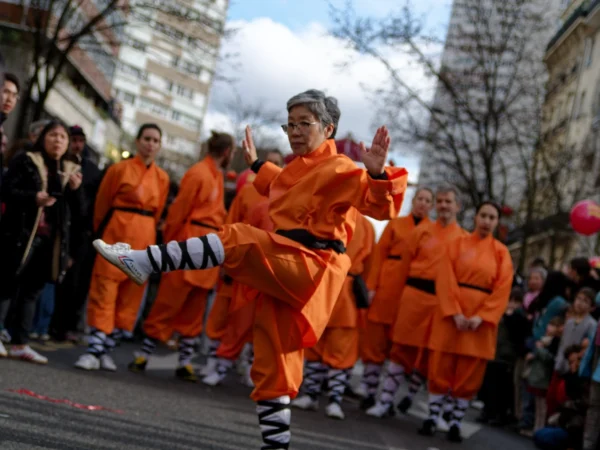 Cyrille · 2026 x 1520 Cette photographie montre un groupe de personnes vêtues de combinaisons orange, participant à une manifestation ou un événement public. Le sujet principal est un individu en mouvement, levant les bras, au centre de l'image.La composition met en valeur une dynamique collective, avec un cadrage serré sur les participants. La lumière est naturelle, probablement en journée, avec des ombres douces. Les couleurs dominantes sont l’orange vif des tenues et des accessoires, contrastant avec les tons neutres de l’arrière-plan urbain.L’ambiance est énergique et militante, évoquant une protestation ou une action symbolique. Le style est documentaire et spontané.