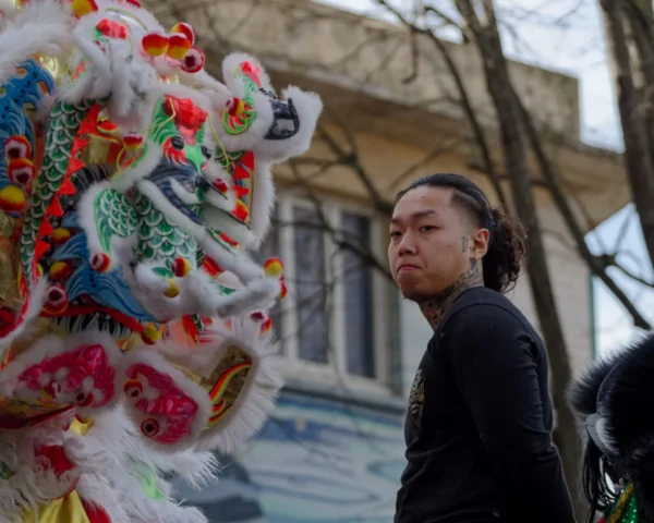 Cyrille · 2026 x 1621 Cette photographie met en scène un jeune homme comme sujet principal, posant devant une tête de lion colorée, typique des défilés traditionnels asiatiques. Le cadrage est serré, centré sur son visage et l'animal, créant une interaction visuelle. La lumière naturelle, douce mais contrastée, accentue les détails et les expressions.Les couleurs dominantes sont vives : rouge, or, bleu et blanc, apportant une ambiance festive et dynamique. Le style est documentaire et spontané, capturant un moment authentique. L’ambiance est à la fois culturelle et intime, mêlant tradition et modernité.