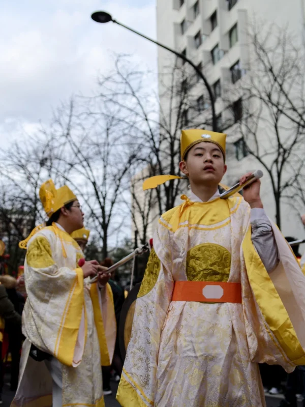Cyrille · 1520 x 2026 Cette photographie montre deux personnes vêtues de costumes traditionnels japonais, probablement des *mikos* ou participants à une cérémonie. Le sujet principal est la personne au premier plan, vêtue de jaune et de blanc, avec une coiffe dorée.La composition est dynamique, avec un cadrage serré sur les personnages. La lumière est naturelle, légèrement diffuse, créant des ombres douces. Les couleurs dominantes sont le jaune vif, le blanc et des touches de rouge et noir.L’ambiance est solennelle et culturelle, évoquant une tradition ou une fête japonaise. Le style est documentaire, mettant en valeur l’authenticité du moment.