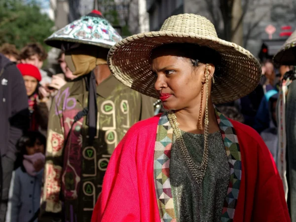 Cyrille · 2026 x 1519 Cette photographie met en scène une personne comme sujet principal, vêtue d’un chapeau de paille, d’un manteau rouge et d’un collier, avec un maquillage rouge aux lèvres. L’arrière-plan montre une foule en costumes colorés, évoquant une fête ou un événement culturel.Le cadrage est serré sur le visage et la partie supérieure du corps. La lumière semble naturelle, bien répartie, mettant en valeur les détails. Les couleurs dominantes sont vives : rouge, vert, et les tons pastel des vêtements.Le style semble documentaire ou artistique, capturant une ambiance festive et traditionnelle. L’atmosphère est joyeuse et animée.