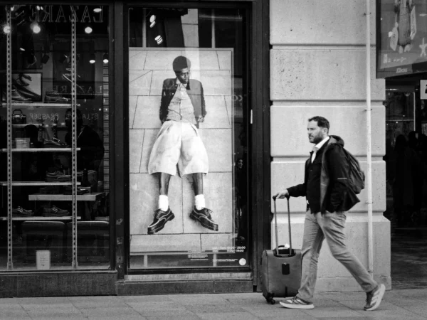 Cyrille GONTIER · 07/03/2025 · 2026 x 1520 Cette photographie en noir et blanc met en scène un homme marchant avec une valise, sujet principal en mouvement. La composition oppose son dynamisme à la statue immobile dans la vitrine, créant un contraste saisissant. Le cadrage serré isole les deux figures, renforçant leur importance.La lumière, douce et naturelle, adoucit les contours et accentue les ombres, ajoutant une touche dramatique. Les tons dominants, du noir au gris clair, renforcent l’ambiance urbaine et mélancolique. Le style est réaliste et documentaire, évoquant une atmosphère contemplative et introspective dans un cadre urbain.